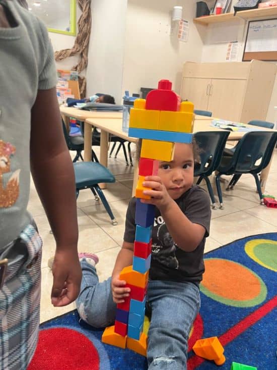 Toddler at Christian daycare in Las Vegas holding up a tall structure made of toy bricks.